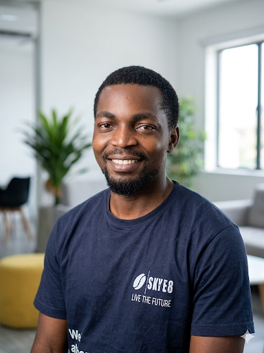 A professional, high-resolution portrait of a confident African male technology executive in a modern, well-lit office setting. He is wearing a dark, branded t-shirt and looking directly at the camera with a warm, encouraging smile. The background is softly blurred, emphasizing depth and a premium corporate environment. Lighting is soft and natural.